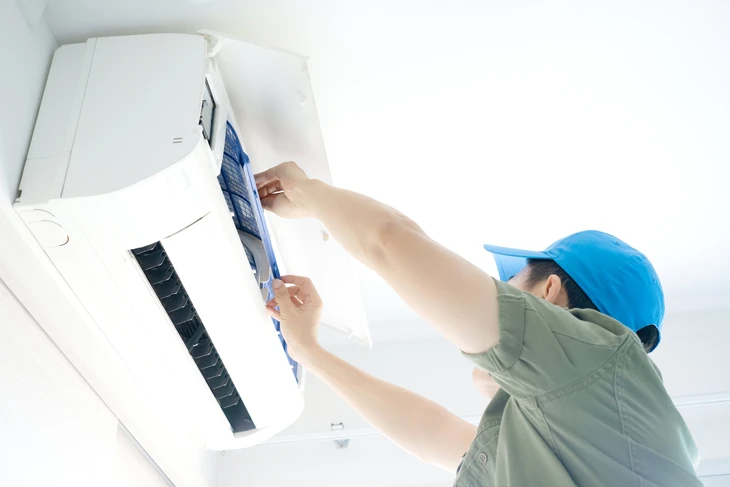 Technicien en salopette bleue effectuant la maintenance d'un climatiseur mural split dans un salon moderne
